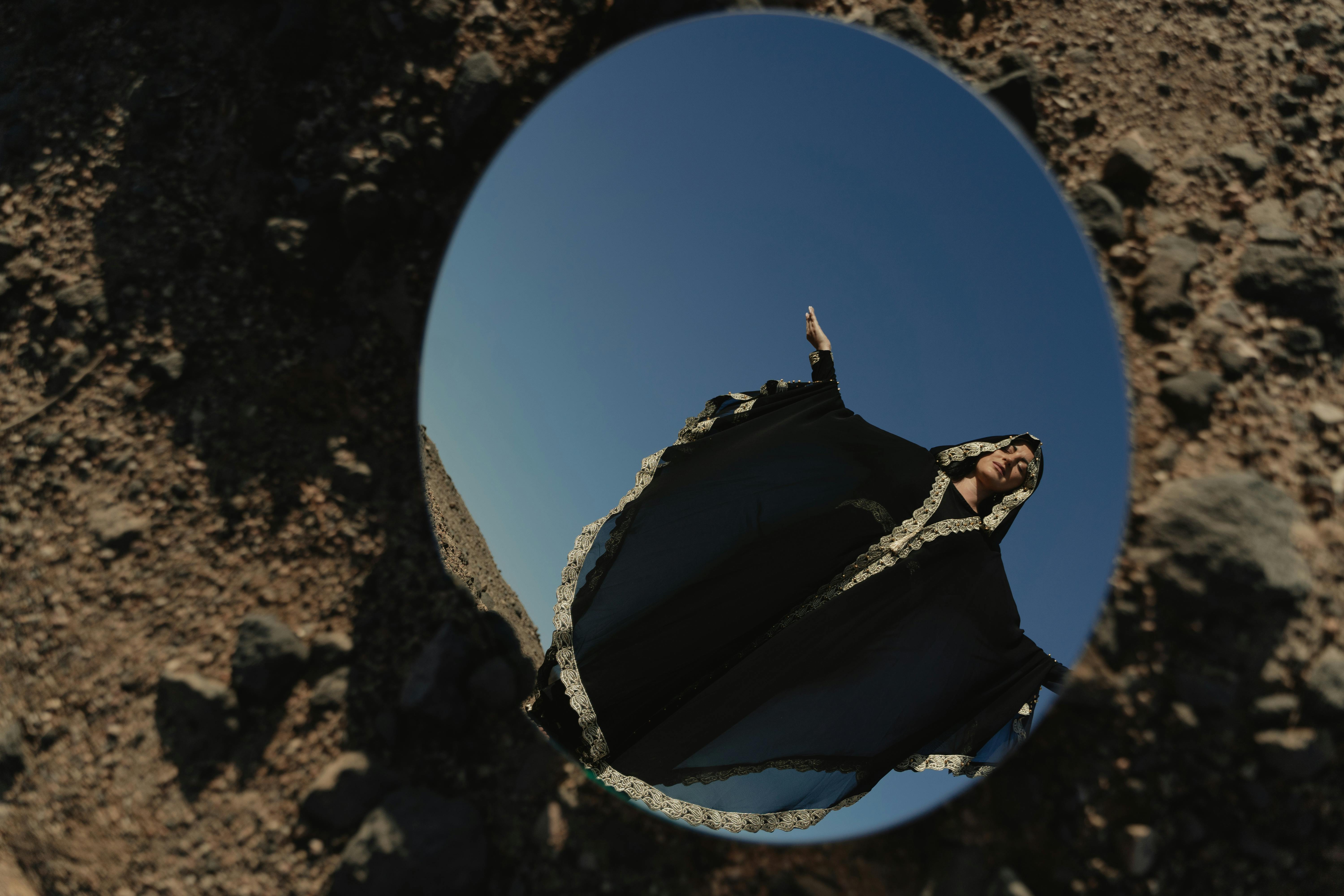 abaya and kandura - A woman in a traditional abaya is reflected in a mirror placed on rocky ground under a clear blue sky.