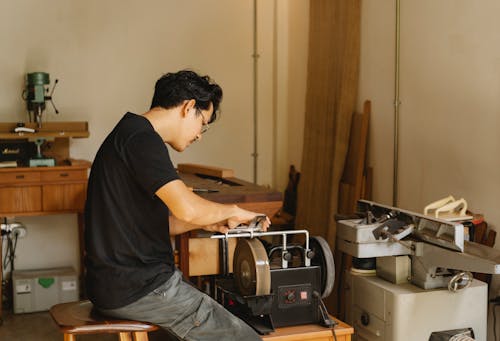 Side view of young concentrated Asian male joiner working on grinder with sharp wheels while sitting on stool in workshop
