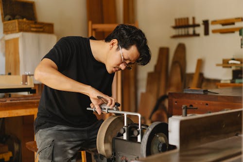 Young attentive ethnic male joiner in eyewear sharpening manual instrument on grinder with wheels in light workroom