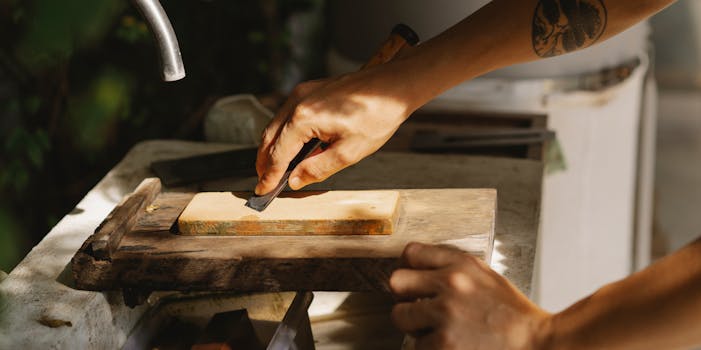 High angle of crop anonymous tattooed woodworker flattening wooden block with sharp tool on old washbasin in sunlight