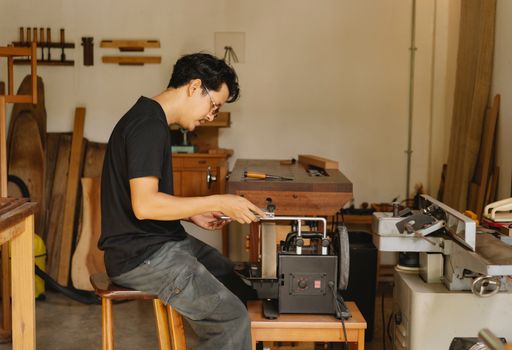 Side view of concentrated skilled ethnic male master sitting at electrical sharpening machine with sharp knife during work in joinery
