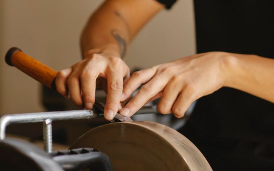 Unrecognizable male carpenter sharpening metal detail on grinding wheel while sitting at machine with metal stick in studio on blurred background