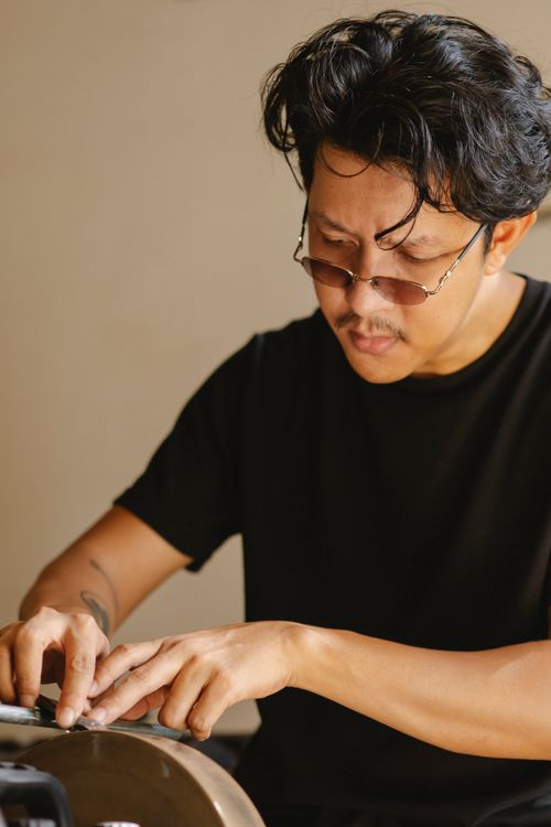 Free Diligent ethnic male master wearing eyeglasses using grinding wheel to sharp knife while working at professional machine against gray wall Stock Photo