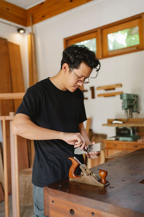 Concentrated male carpenter in eyeglasses standing with metal knife at wooden workbench with jointer during work process in joinery workshop