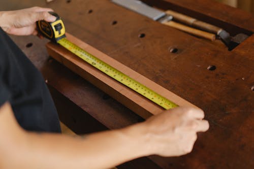 Free Unrecognizable male carpenter standing at wooden workbench and measuring length of board with yellow tape measure while working in professional studio Stock Photo