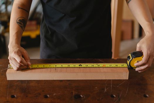 Unrecognizable male carpenter in black outfit with tattoo using ruler on wooden plank while working in studio on blurred background