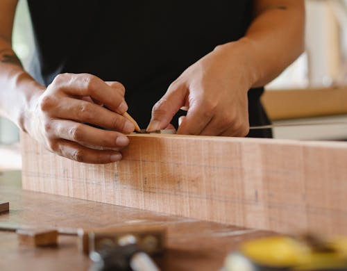 Free Unrecognizable male master using pencil and ruler to measure width of wooden detail while working in professional workshop on blurred background Stock Photo