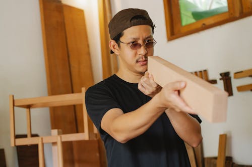 Pensive attentive ethnic male in eyeglasses checking flatness of timber plank in workshop in daytime