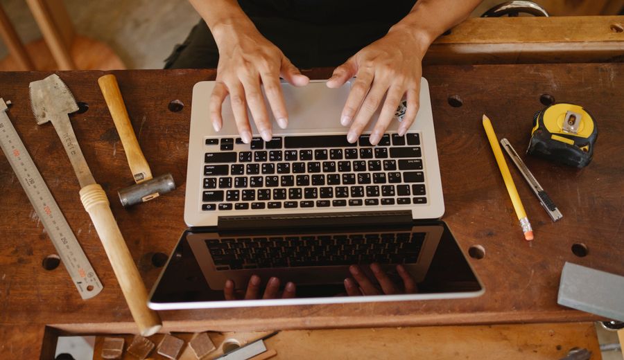Dłonie piszące na laptopie na stole warsztatowym obok młotka, dłuta, linijki, ołówka i miarki