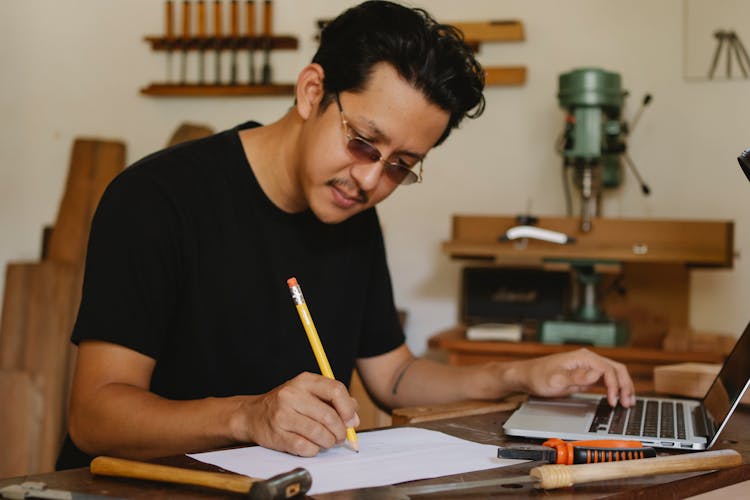 Thoughtful Man Writing Notes While Working In Carpenter Studio