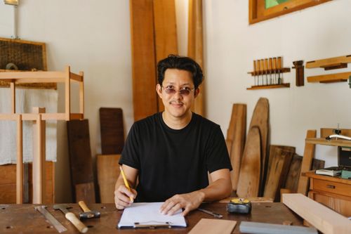 Free Cheerful ethnic male in casual clothes and eyeglasses sitting at table with instruments while working with new project and looking at camera Stock Photo