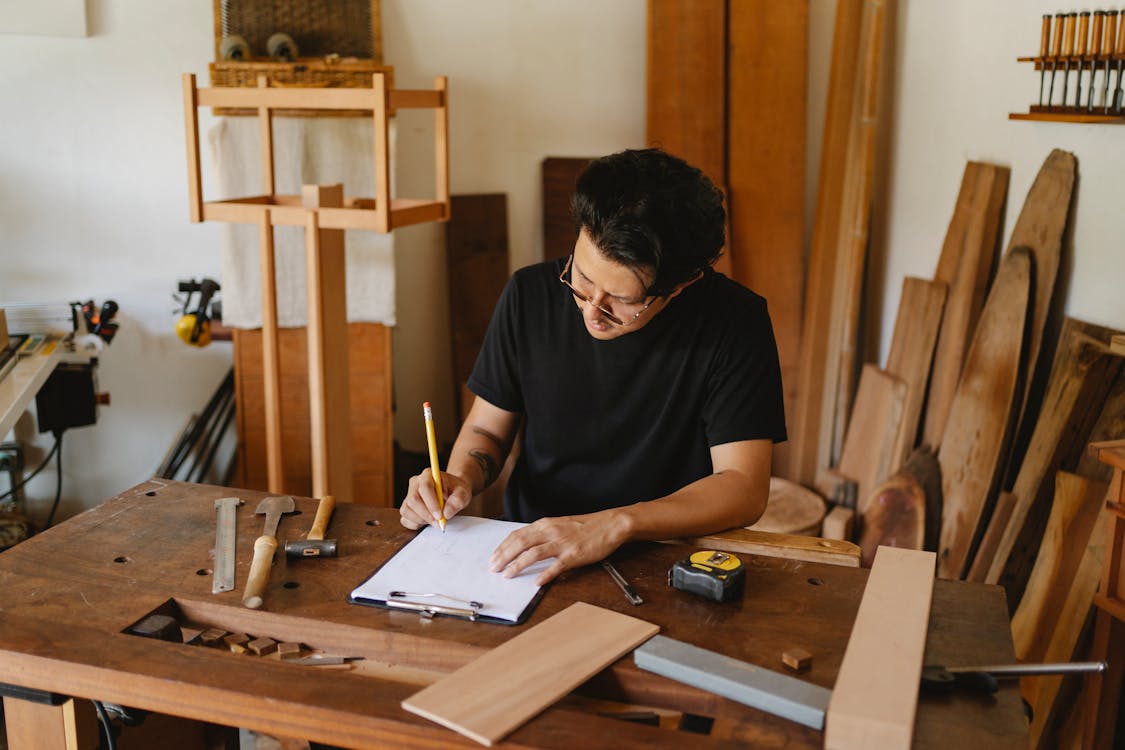 Free Concentrated man in glasses sitting at wooden table in workshop and holding pencil while creating new object on paper Stock Photo