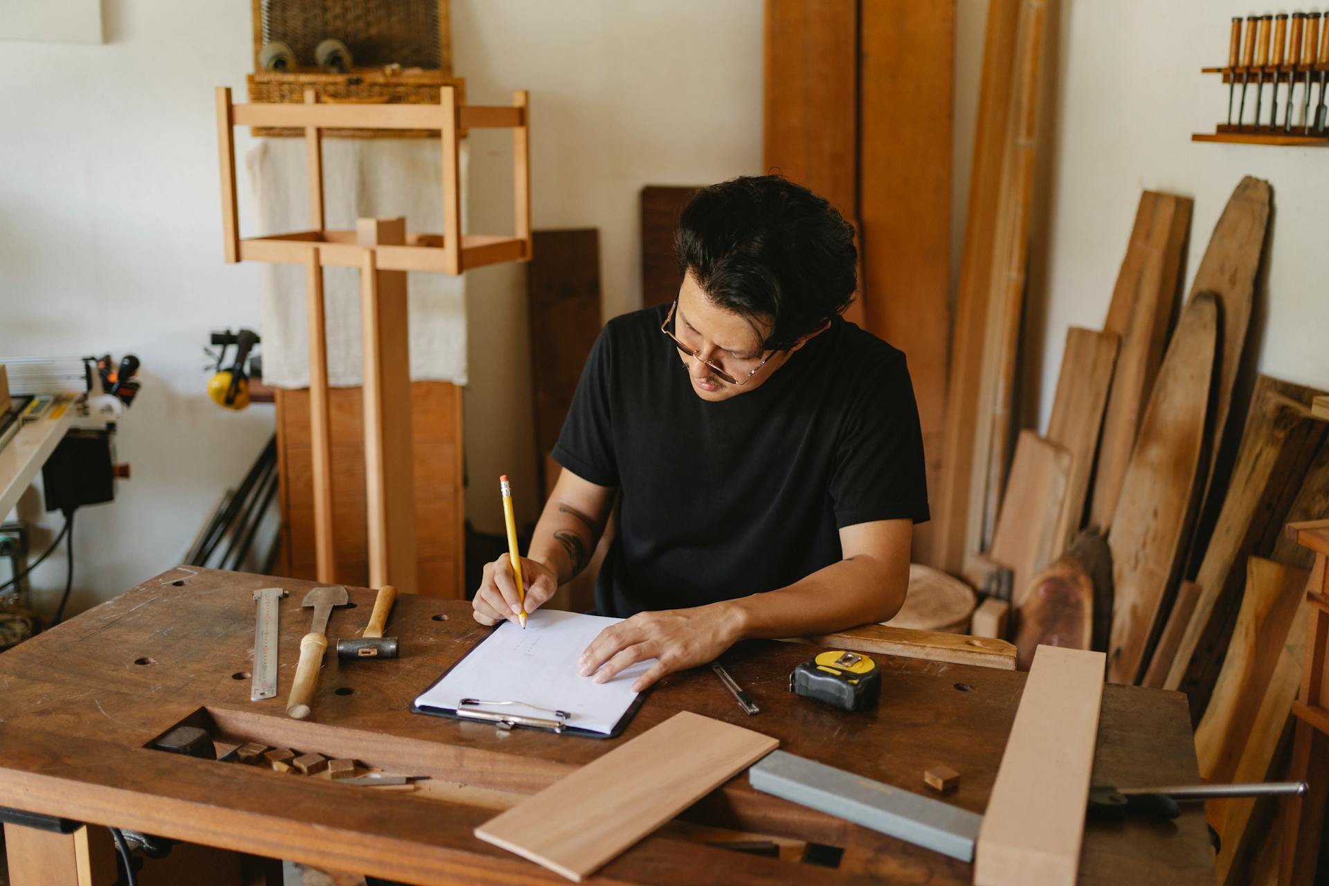 Concentrated man in glasses sitting at wooden table in workshop and holding pencil while creating new object on paper