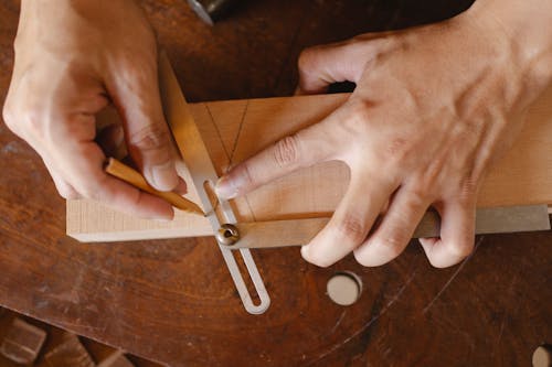 From above of crop anonymous craftsman using pencil and bevel gauge while working with wooden detail