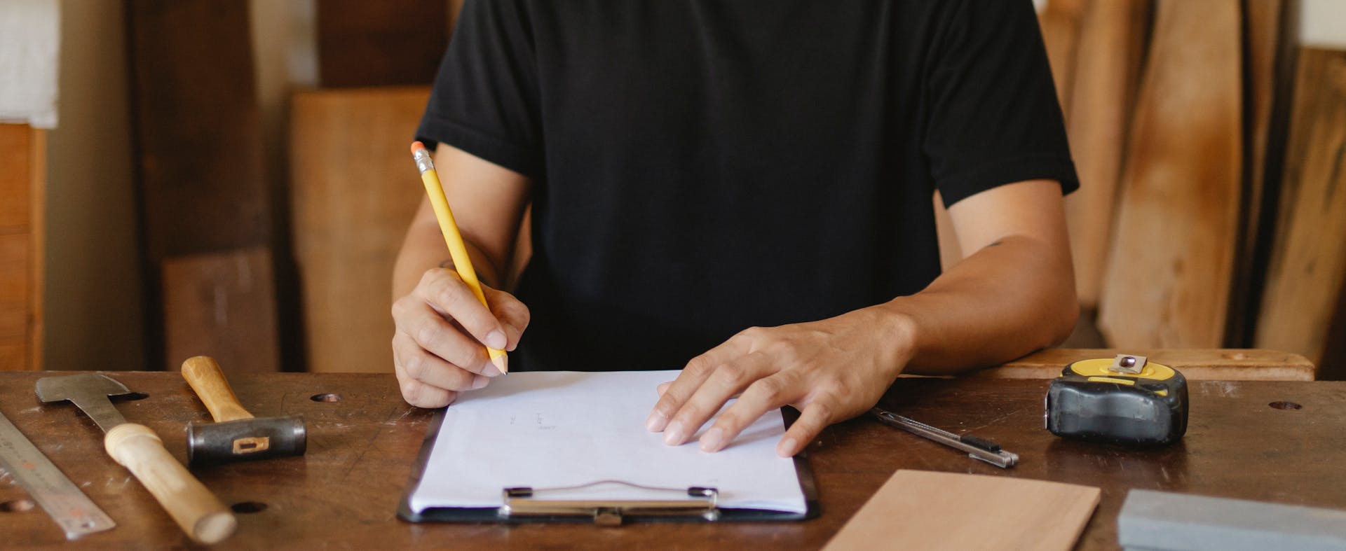 Crop anonymous man sitting at wooden table with instruments and holding pencil while sketching new project on paper