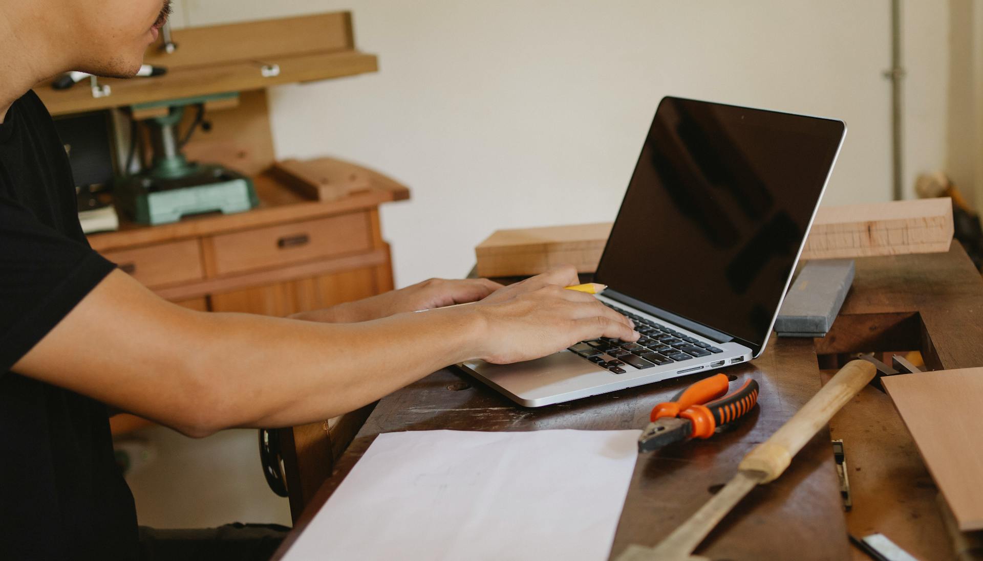Craftsman working on a laptop in a woodwork shop with tools and paper plans around.