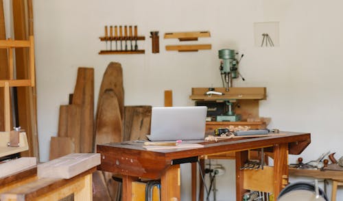 Workroom interior with assorted wooden pieces near wall and drill press against netbook in daylight