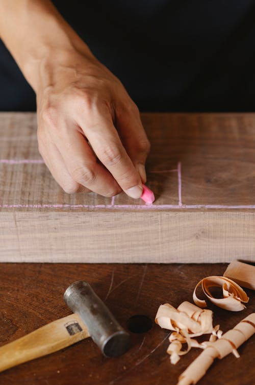 From above of crop anonymous joiner drawing vertical line on wooden block with colored chalk on table with shavings and mallet