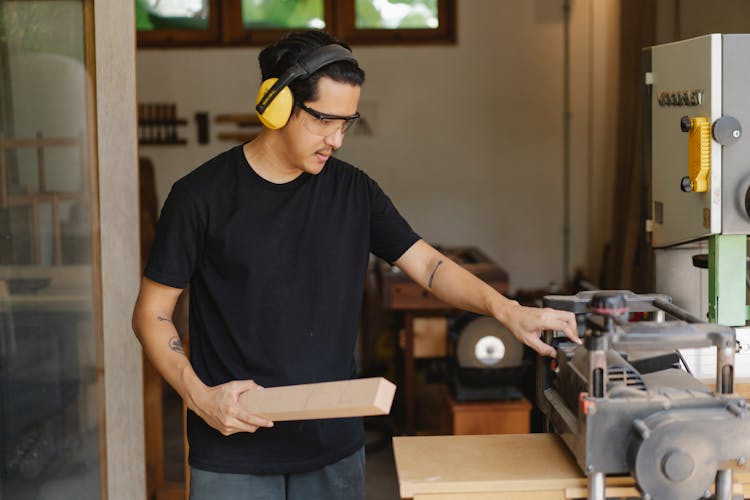 Asian Carpenter With Lumber Near Planer And Workshop