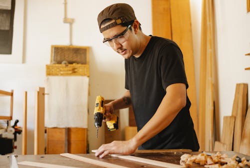 Young attentive ethnic male carpenter in protective glasses with drilling machine and timber in workroom