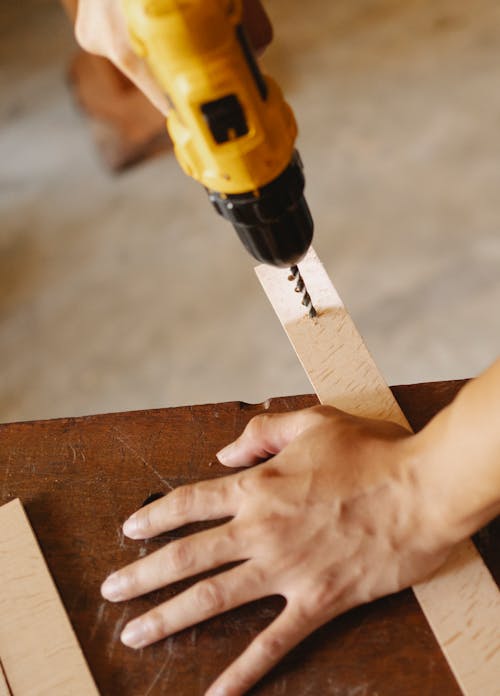 Free Crop unrecognizable carpenter using drill to create hole in wooden board while working in workshop Stock Photo