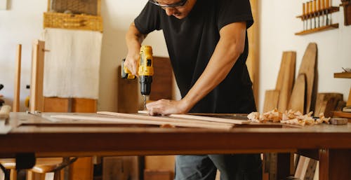 Crop faceless man standing near table and making hole with drill while working in workshop