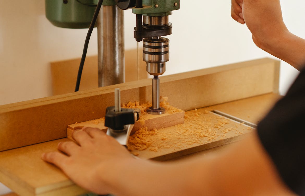 Crop anonymous carpenter using drill press to cut wooden detail while creating new furniture in workshop