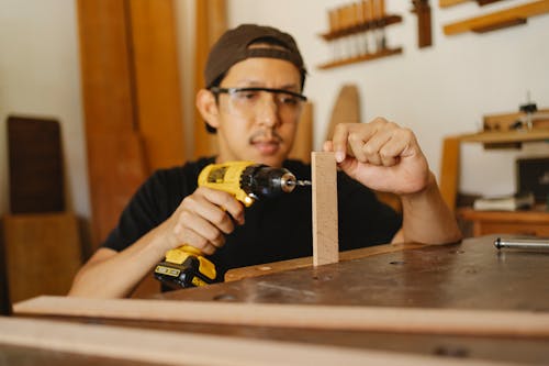 Focused Asian male in cap and glasses holding drill and wooden detail while working in modern workshop