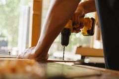Crop anonymous male drilling timber plank with professional screwdriver on table in workshop