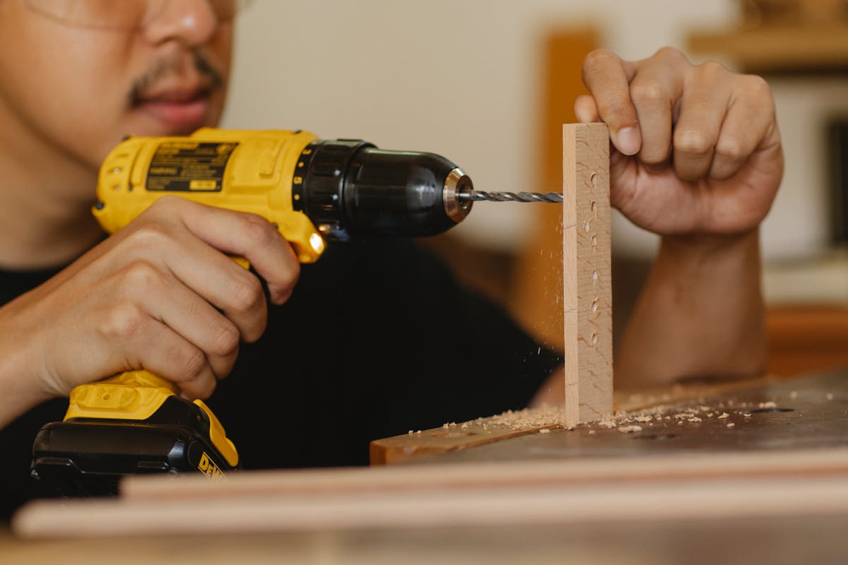 Crop anonymous male drilling holes in wooden panel while working on table in workshop