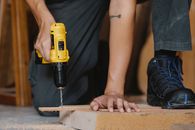 Crop anonymous male drilling and pressing timber detail with foot in boot in workshop
