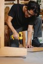 Focused ethnic male in safety glasses drilling wooden plank with screwdriver among lumber in workshop