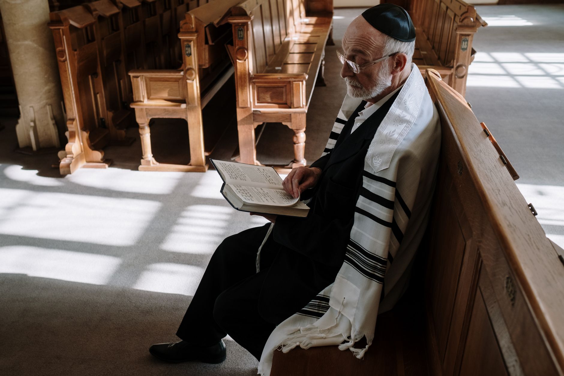Elderly man wearing kippah and tallit, reading in a synagogue. Traditional Jewish setting.