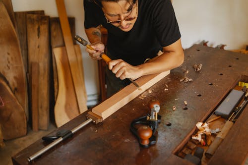 Free High angle of crop craftsman in eyeglasses cutting wooden detail with hammer and chisel while working in joinery Stock Photo