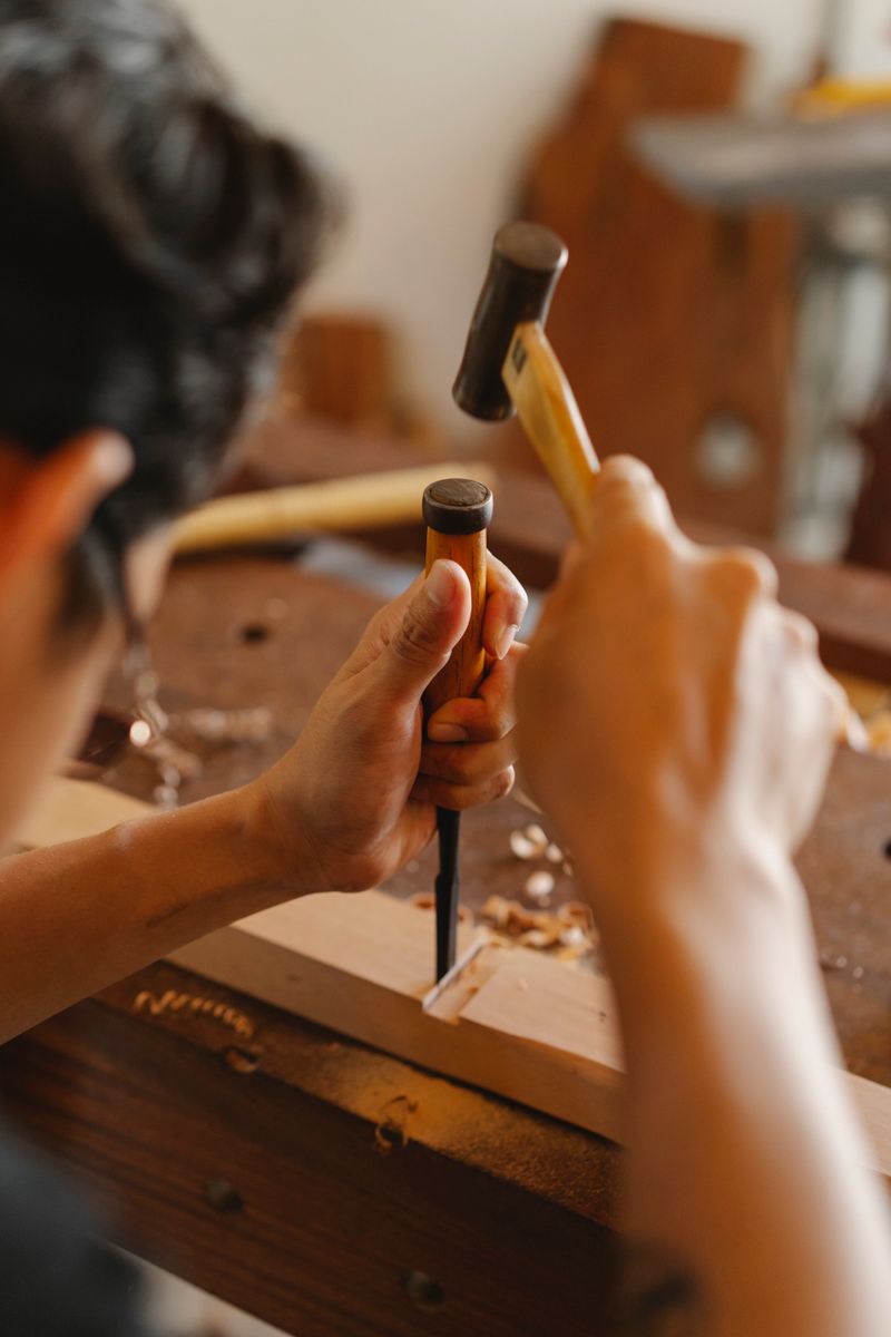 Crop anonymous carpenter in glasses forming wooden detail with hammer and chisel while working in joinery