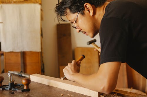 Side view of concentrated Asian male master in eyeglasses processing wooden plank with chisel and hammer in workroom