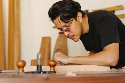 Concentrated precise Asian craftsman in eyeglasses creating wooden detail with chisel while working in joinery