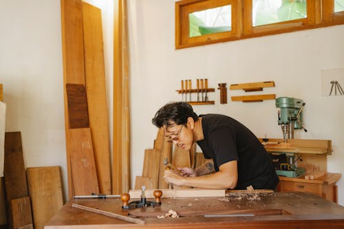 Side view of concentrated Asian craftsman creating wooden plank with hammer and chisel during work in professional joinery