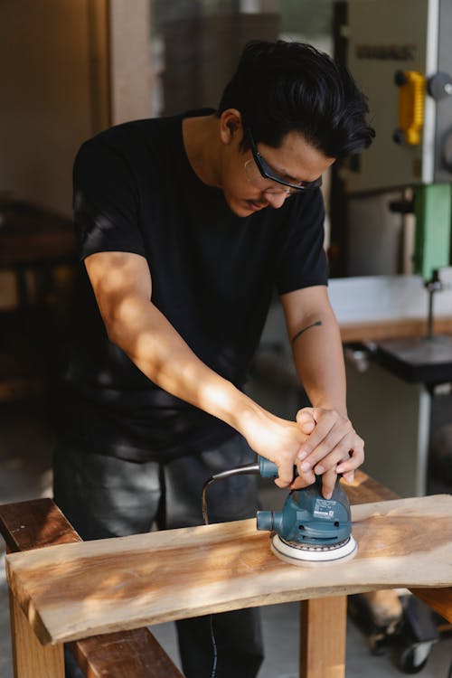 Concentrated male woodworker in protective goggles standing in carpentry workshop and polishing wooden object with sander