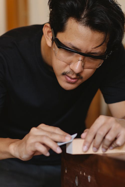 Free Concentrated skilled craftsman in glasses correcting form of wooden plank with sandpaper while working in carpentry workshop Stock Photo