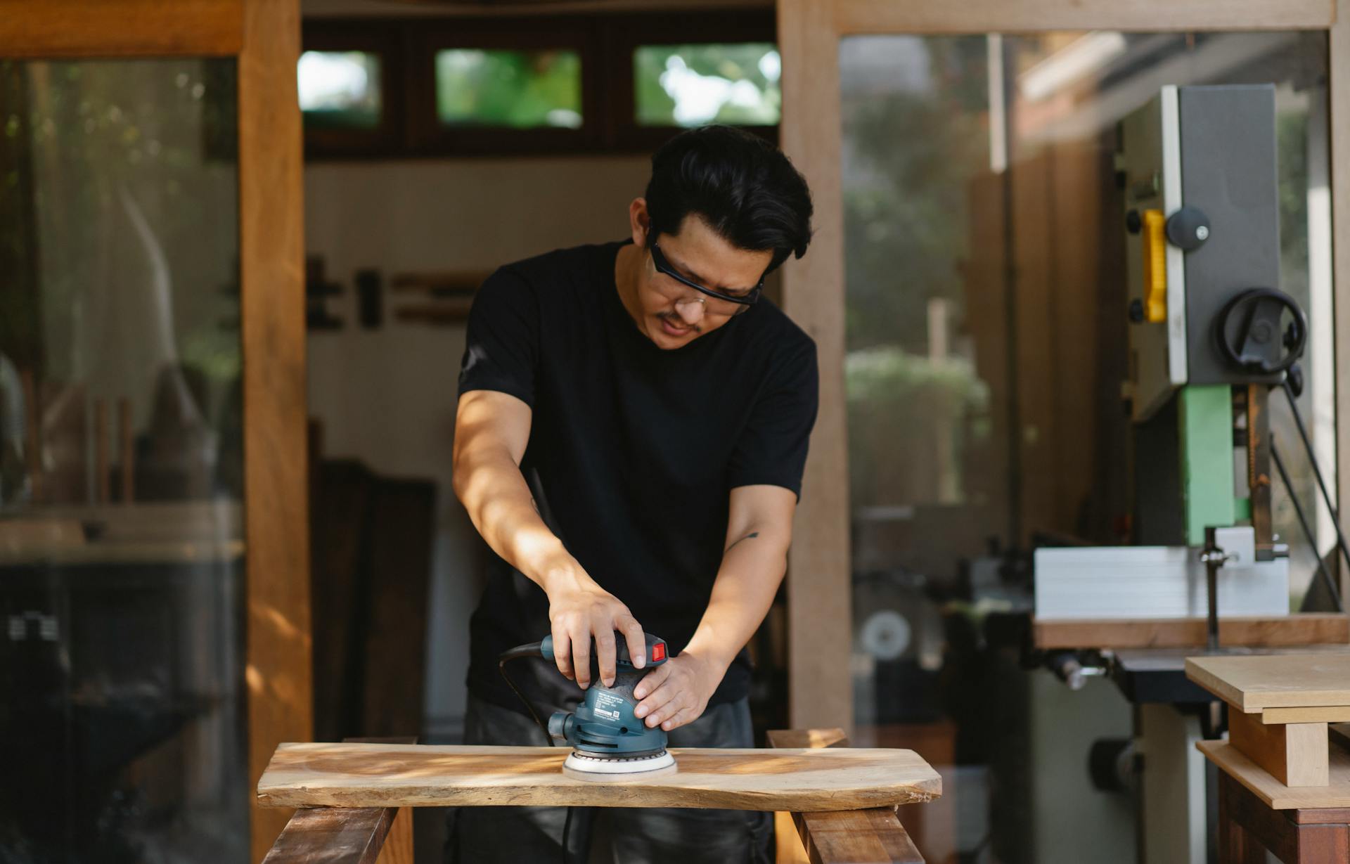 Concentrated male master in protective glasses smoothing wooden plank with sander while creating wooden detail in workroom