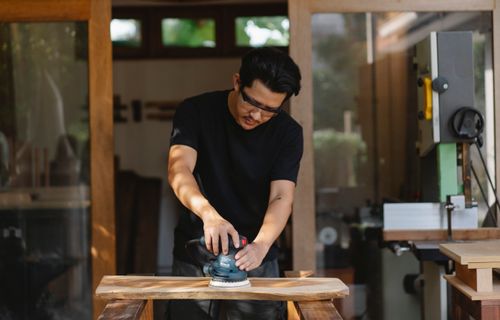 Free Concentrated male master in protective glasses smoothing wooden plank with sander while creating wooden detail in workroom Stock Photo