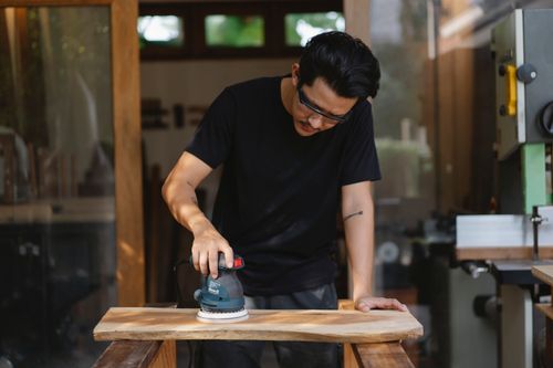 Free Concentrated male woodworker in glasses polishing wooden plank with sander while making lumber detail in joinery Stock Photo