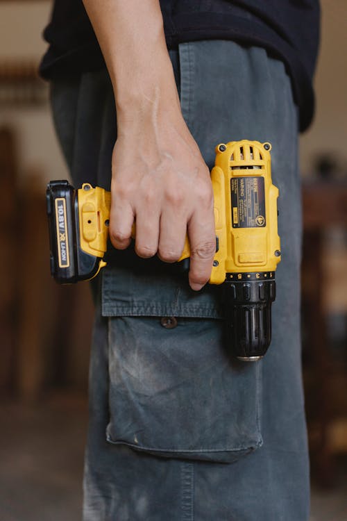Side view of unrecognizable craftsman in casual clothes standing with electric cordless screwdriver in workshop