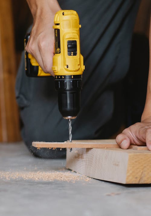 Crop unrecognizable carpenter in black outfit using modern yellow screwdriver to drill hole in wooden blank while working in joinery