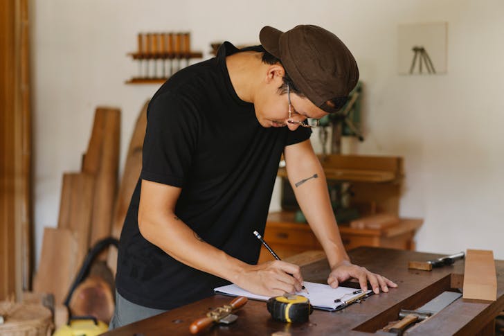Side view of Asian man in casual clothes and eyeglasses bending on table and taking notes by pencil while working in joinery