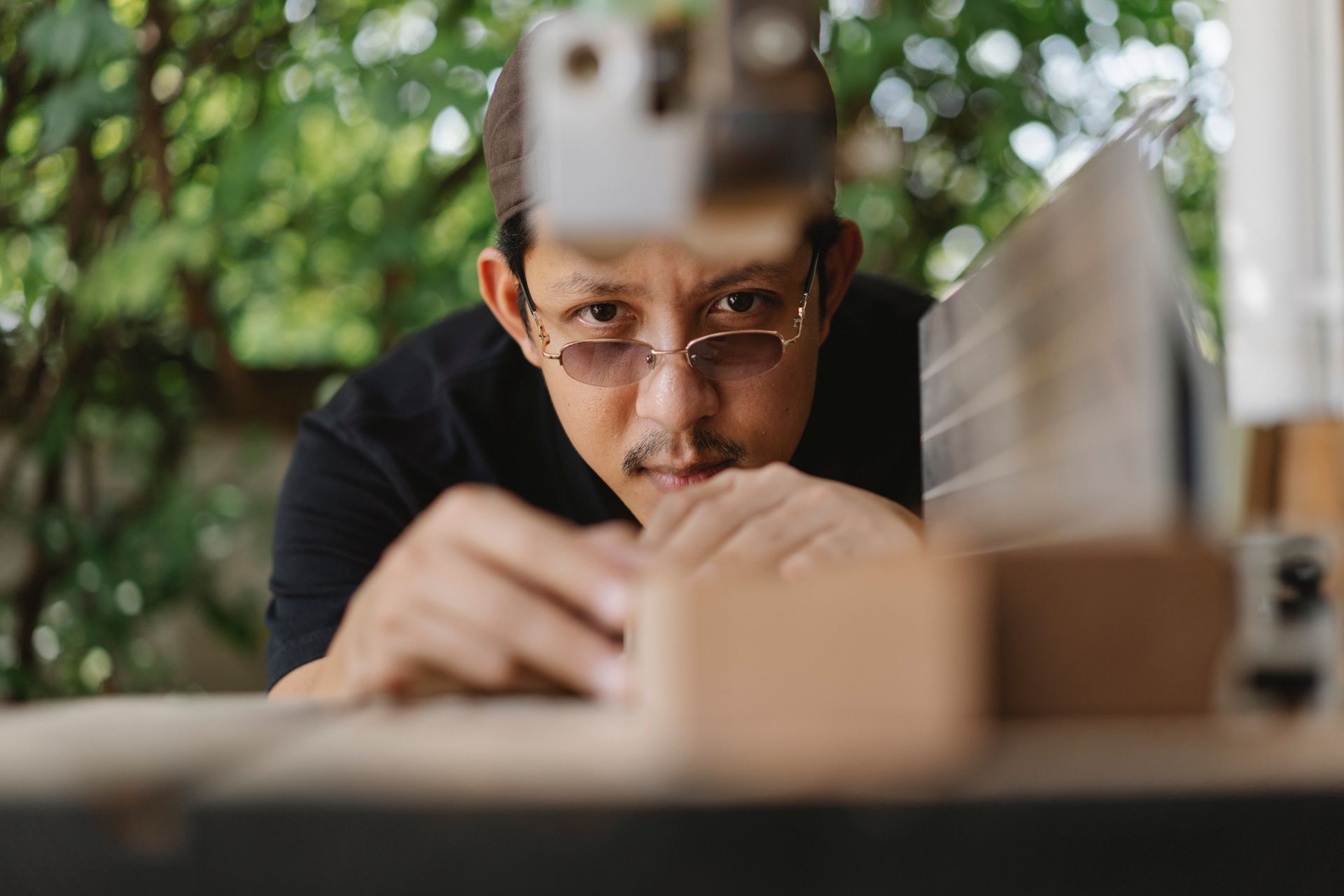 Concentrated ethnic craftsman in casual wear in hat and eyeglasses working on workbench with wood plank outside near workshop at daytime