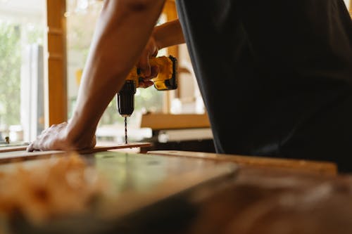 Unrecognizable guy drilling wooden board in workshop