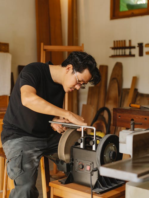 Side view of focused young ethnic male carpenter in casual clothes and eyeglasses using pedestal grinder while creating wooden object in workshop