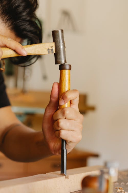 Crop man carving wooden plank with chisel and hammer in workshop